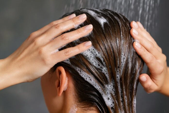 Nahaufnahme einer Frau beim Haarewaschen unter der Dusche, Hände massieren Shampoo-Schaum ins nasse Haar – Symbolfoto für Haarpflege. Nahaufnahme einer Frau beim Haarewaschen unter der Dusche, Hände massieren Shampoo-Schaum ins nasse Haar – Symbolfoto für Haarpflege.